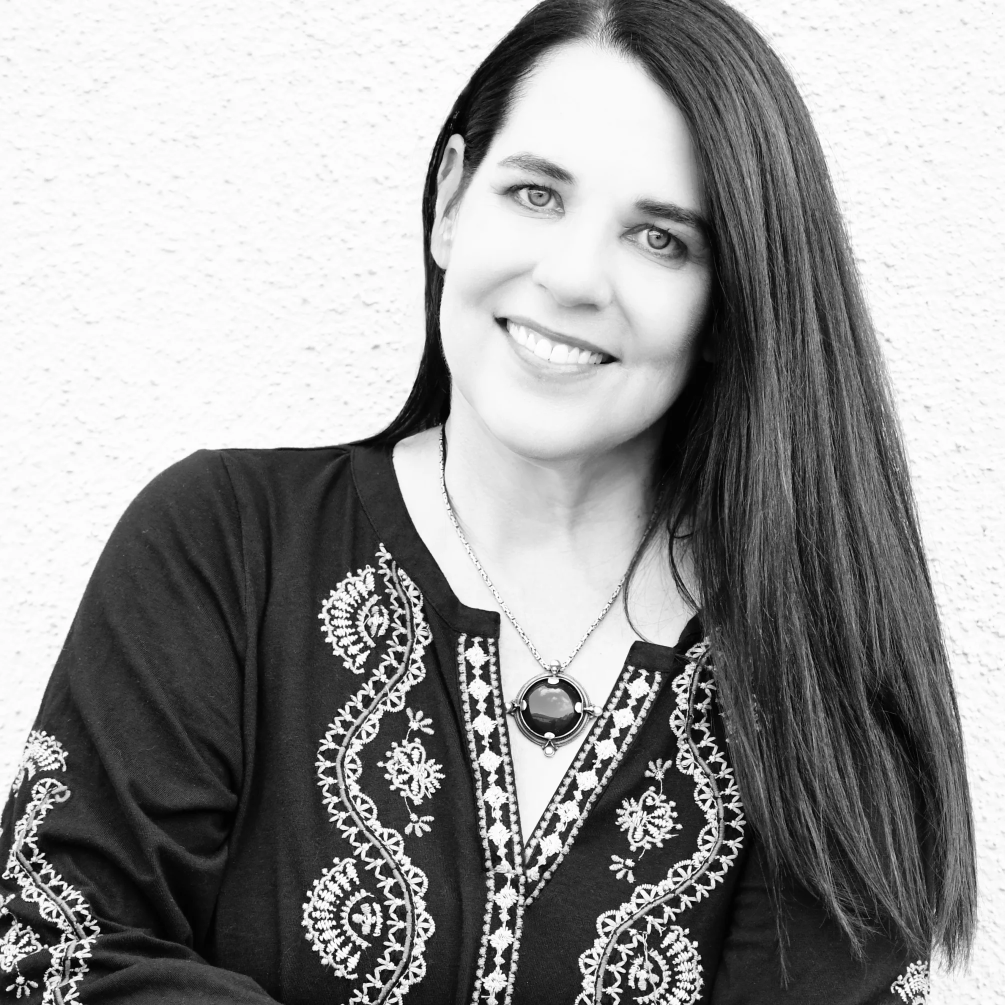 A black and white image of the CEO of Jackrabbit, Mary Maltbie, a smiling woman with long dark hair, necklace, dark blouse with detailed patterns along the seams, on a white background.